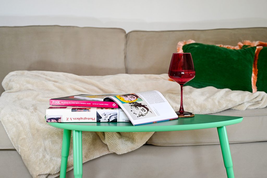 A close-up of a cozy beige sofa with a deep red wine glass and design books on a mint green side table, creating a stylish, relaxed setting for reading and unwinding.