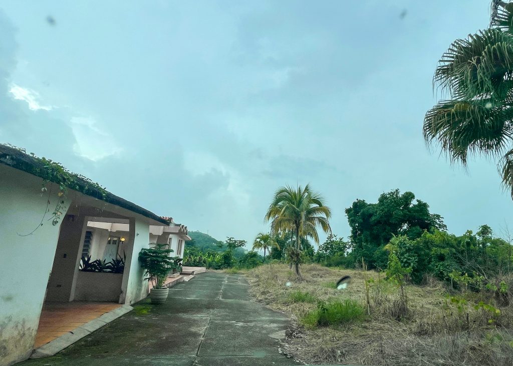 Original state of Hacienda Eterna Primavera, with weathered facade, overgrown lawn, and a cloudy tropical sky in the background.